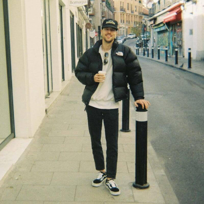 Der Berufswahl-Coach, Simon Der Berufswahl-Coach, Simon, steht auf einer Strasse mit einem Kaffee in der Hand und lächelt in die Kamera